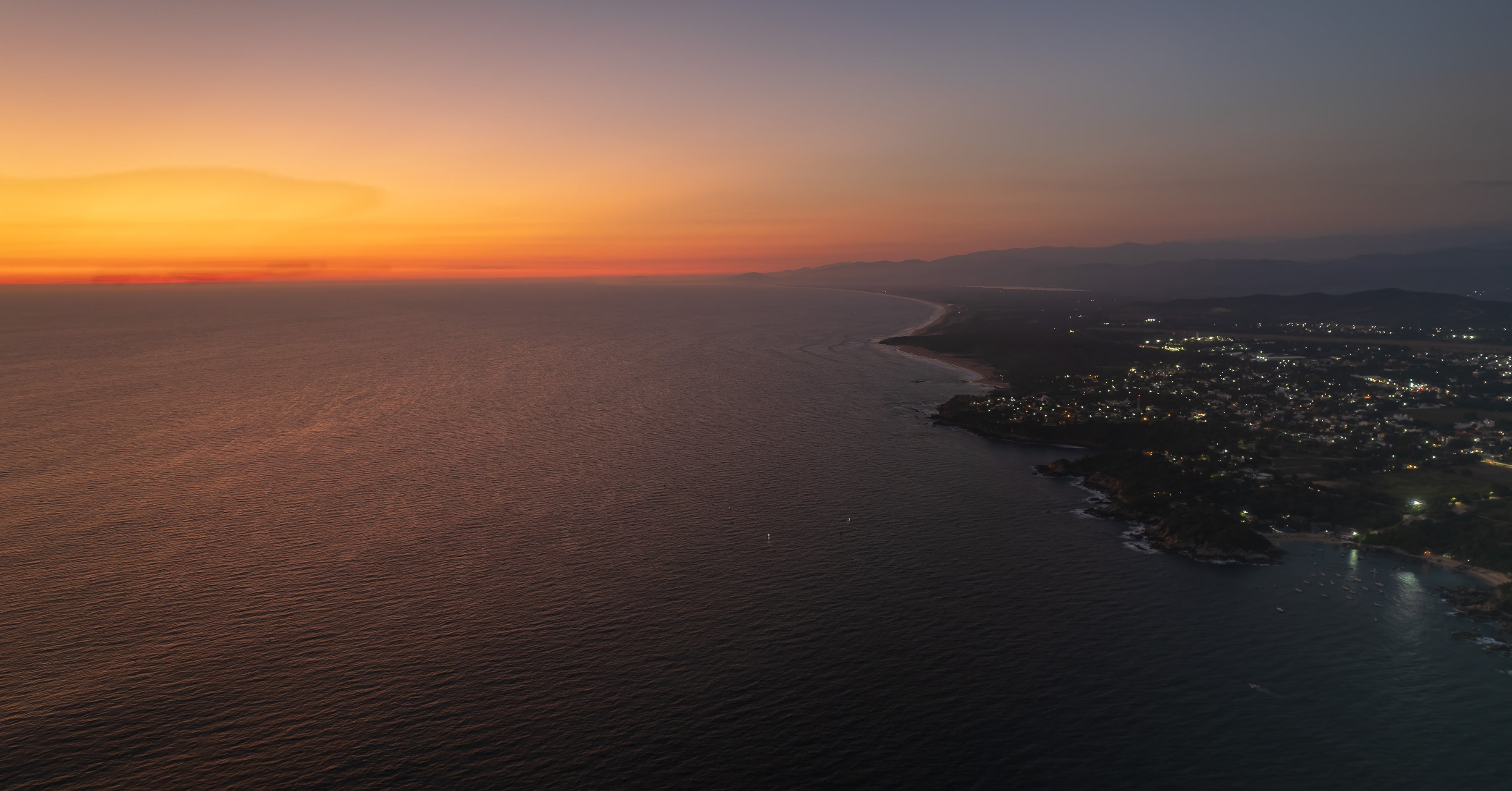 Aerial view of a coastal city at sunset with orange sky and dark water.